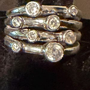 May include: A stack of four silver-toned rings, each featuring a textured, organic band. Each ring is set with a single round, clear, faceted stone in a bezel setting. The stones vary in size across the four rings, creating a layered appearance.