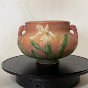 May include: A ceramic vase with a peach-colored exterior and a white and green floral design. The vase has two handles and is sitting on a black rotating stand. The floral design includes a white flower and green leaves.