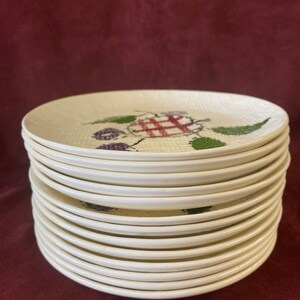 May include: A stack of 15 white ceramic plates with a red and green berry design. The plates are stacked neatly on a burgundy surface.