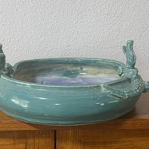 May include: A teal ceramic bowl with two frog-shaped handles. The bowl has a light blue and white glaze inside.