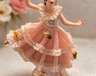 May include: A porcelain figurine of a dancer in a pink dress with intricate lace-like texture, ruffled layers, and small yellow flower accents. The figure has brown hair, red lips, and is posed with arms extended, standing on a gold-trimmed base.