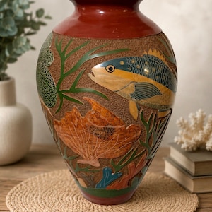 May include: A large, bulbous ceramic vase with a smooth, dark red rim and base. The textured, brown body features intricate, colorful relief carvings of a fish, coral, and seaweed in shades of yellow, blue, green, and orange, creating a detailed underwater scene.