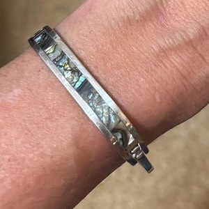 May include: A silver bracelet with an abalone shell inlay. The bracelet is rectangular with a hinged clasp. The abalone shell has iridescent colors of blue, green, and silver. The bracelet is worn on a wrist.