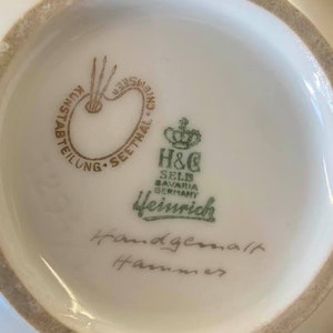 May include: Close-up of the bottom of a white porcelain vase with a green and gold logo that reads "H&C Selb Bavaria Germany Heinrich" and the words "Handgemalt Hammer" in cursive handwriting.