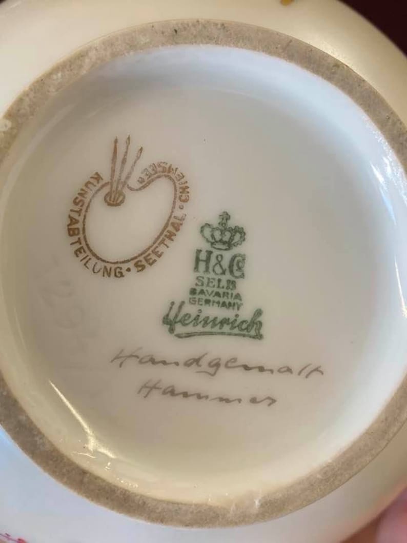 May include: Close-up of the bottom of a white porcelain vase with a green and gold logo that reads "H&C Selb Bavaria Germany Heinrich" and the words "Handgemalt Hammer" in cursive handwriting.
