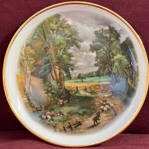 May include: A white ceramic plate with a gold rim features a vintage illustration of a rural landscape with trees, a path, and sheep. The scene is painted in shades of green, brown, and blue.
