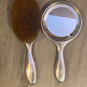 Puede incluir: Un espejo de mano plateado y un cepillo para el cabello con cerdas marrones. Ambos tienen un mango plateado.
