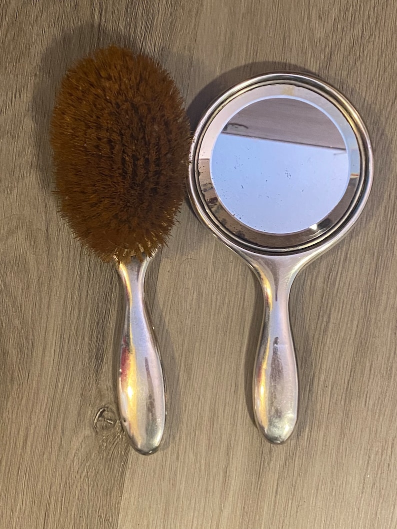 Puede incluir: Un espejo de mano plateado y un cepillo para el cabello con cerdas marrones. Ambos tienen un mango plateado.