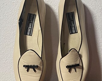 May include: A pair of off-white leather loafers with black trim and a black bow detail. The loafers are by Stuart Weitzman, New York.