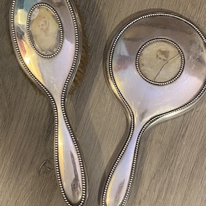 Puede incluir: Un juego de espejo de mano y cepillo para el cabello plateado con un borde de cuentas. Cada pieza presenta un inserto circular con una imagen descolorida de un ni&ntilde;o.