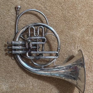 May include: A silver French horn with intricate tubing and a flared bell. The bell features an etched floral design. The instrument is designed for playing music and is made of shiny metal.
