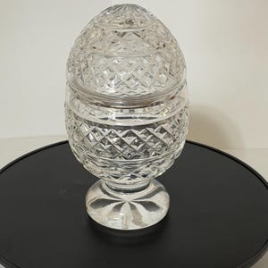 May include: A clear, cut-glass egg-shaped jar with a lid. The jar features a diamond pattern and sits on a round, clear base. The jar is displayed on a black surface.
