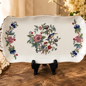 May include: A rectangular porcelain serving tray with a floral and bird design. The tray features a white background with a central bird and floral motif in pink, blue, and red. The edges are scalloped and trimmed with gold.