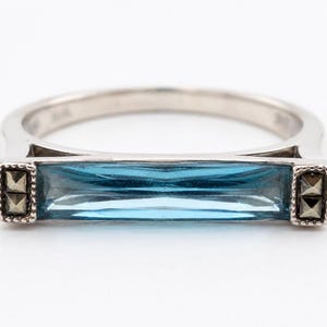 May include: A silver-toned ring featuring a long, rectangular, baguette-cut blue gemstone set horizontally. The stone is flanked on both sides by small, square, dark metallic accents with a pyramid-like texture, all set within a polished metal band.