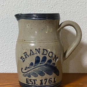 May include: A gray stoneware pitcher with a blue and white design. The pitcher has a handle and says "Brandon Est. 1761" on the front.