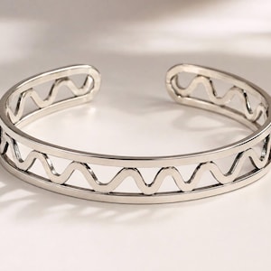 May include: A silver-toned open cuff bracelet featuring a continuous horizontal zigzag pattern cut out between two smooth, solid metal bands. The piece has a polished, reflective finish and a wide gap at the back, allowing for an adjustable fit.