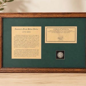 May include: A framed display featuring a silver coin, a Certificate of Authenticity, and a historical information card titled America's First Silver Dollar 1772-1825. The display is set against a dark green mat inside a rectangular wooden frame.