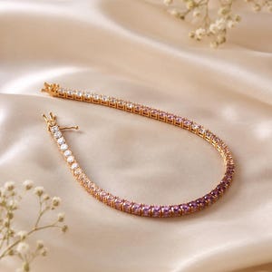 May include: A gold-tone tennis bracelet featuring a gradient of round-cut stones that transition in color from clear white to light pink, lavender, and deep purple. The bracelet includes a secure metal box clasp closure at one end.