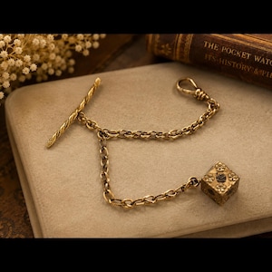 May include: A gold-toned pocket watch chain with a T-bar, chain links, and a decorative square pendant. The pendant has floral designs and a dark blue center. The chain is displayed on a neutral surface.