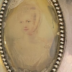 Puede incluir: Un medall&oacute;n ovalado de plata con un borde de cuentas. El medall&oacute;n contiene un retrato en miniatura de una mujer con cabello rubio, vestida con un vestido blanco. El retrato est&aacute; descolorido y tiene un aspecto vintage.