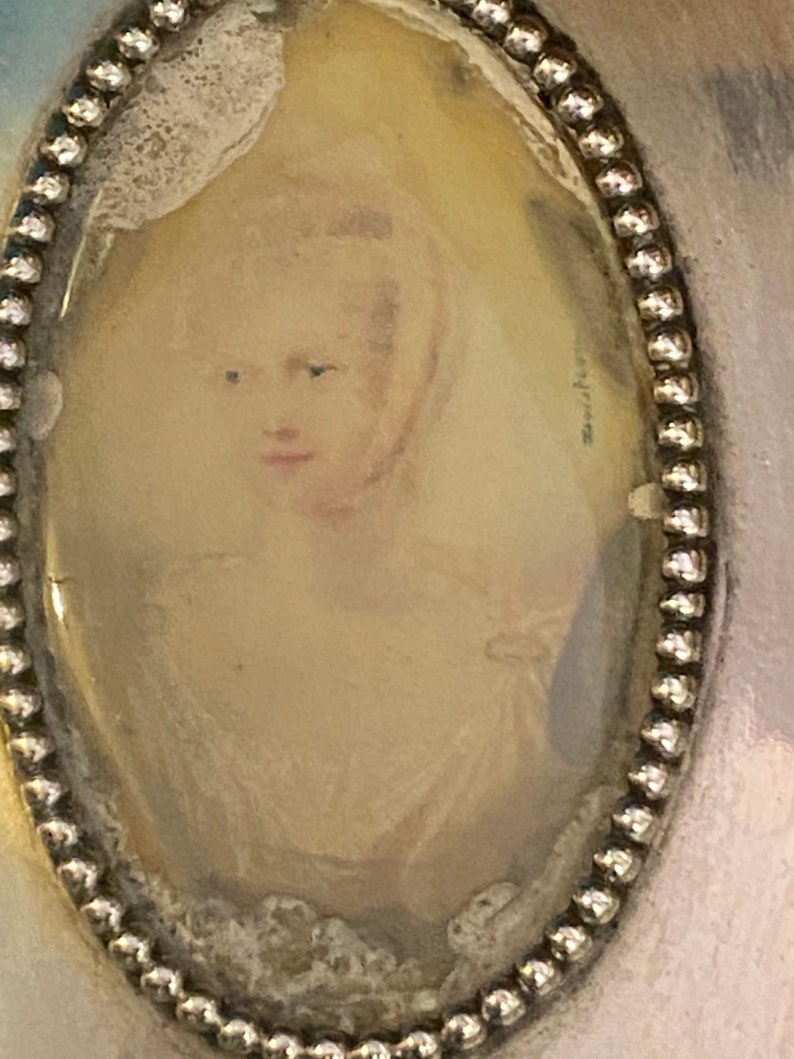 Puede incluir: Un medall&oacute;n ovalado de plata con un borde de cuentas. El medall&oacute;n contiene un retrato en miniatura de una mujer con cabello rubio, vestida con un vestido blanco. El retrato est&aacute; descolorido y tiene un aspecto vintage.