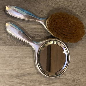 Puede incluir: Un espejo de mano plateado y un cepillo para el cabello plateado con cerdas marrones. El espejo tiene una forma redonda y un mango plateado. El cepillo tiene una forma redonda y un mango plateado.