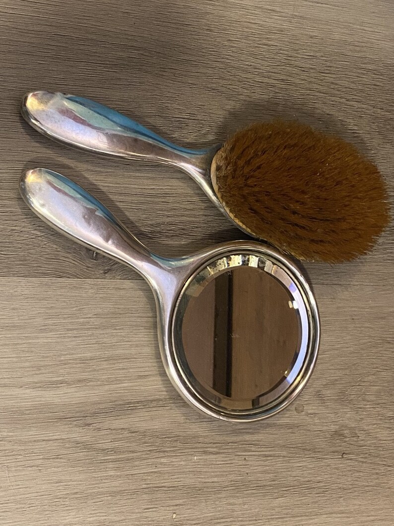 Puede incluir: Un espejo de mano plateado y un cepillo para el cabello plateado con cerdas marrones. El espejo tiene una forma redonda y un mango plateado. El cepillo tiene una forma redonda y un mango plateado.