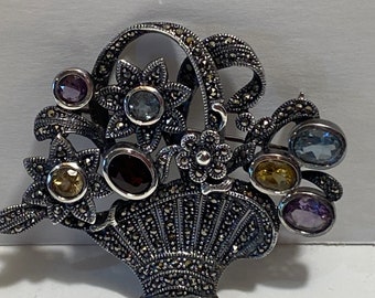 May include: A silver brooch with a basket design, featuring a bouquet of flowers with colourful gemstones. The flowers are made of marcasite and the gemstones are amethyst, topaz, and citrine.