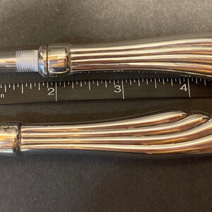 May include: Two silver makeup brushes with a decorative, fluted handle. The brush on the left has a clear plastic tube and a small, fine brush head. The brush on the right has a brown, fluffy brush head.