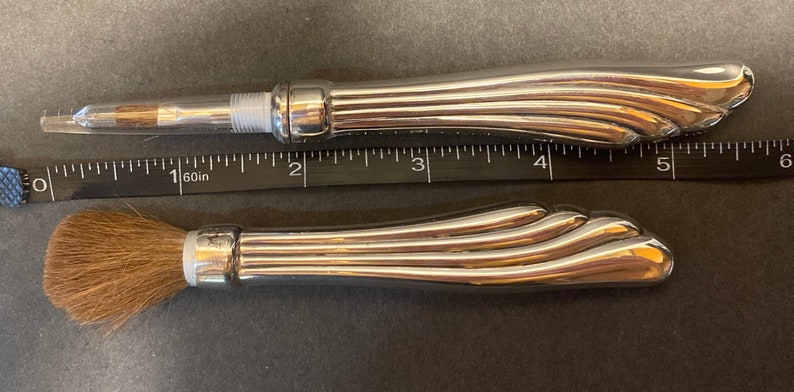 May include: Two silver makeup brushes with a decorative, fluted handle. The brush on the left has a clear plastic tube and a small, fine brush head. The brush on the right has a brown, fluffy brush head.