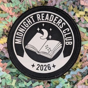 May include: A circular black and white patch featuring the words "MIDNIGHT READERS CLUB" around a graphic of an open book, a crescent moon, and stars. The year "2026" is below the book graphic. The patch is set against a colourful shredded paper background.