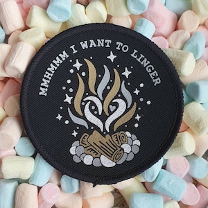 May include: Black round patch with white text that reads "Mmmmmm I want to linger" and a white and gold illustration of a campfire with stars. The patch is surrounded by pastel-coloured marshmallows.