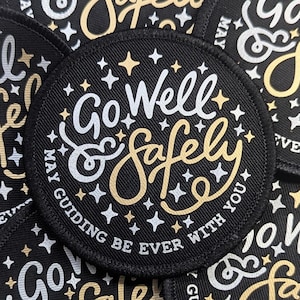 May include: Circular black patch embroidered with the words "Go Well & Safely" in white and gold script, with star accents. The phrase "May guiding be ever with you" is also included. The patch has a black border and is likely an embroidered design.