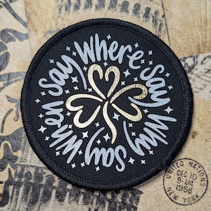 May include: A black round patch with a white and gold design. The design features a four-leaf clover with the words "Where's my son" written around it.