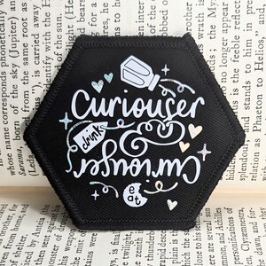 May include: A black hexagonal patch with white text that reads "Curiouser and Curiouser" with a small white image of a bottle and a tag that says "drink". The patch has a glittery, iridescent finish.
