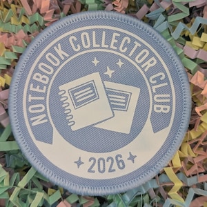 May include: A circular, pale blue patch featuring the text "NOTEBOOK COLLECTOR CLUB". Two white notebook illustrations are in the centre. The year "2026" is at the bottom, with star accents. The patch has a blue stitched border and is surrounded by colourful confetti.