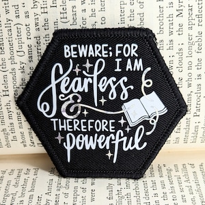 May include: A black hexagonal patch with white text and a white image of an open book. The text reads "Beware: For I am fearless & therefore powerful".