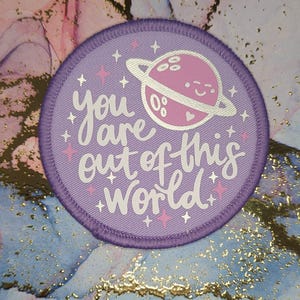 以下が含まれることがあります： 「You are out of this world」という白い文字が書かれた、丸い紫色のパッチ。リングと笑顔の顔を持つピンク色の惑星が星に囲まれています。パッチはアイロン接着または縫い付けのパッチである可能性があります。