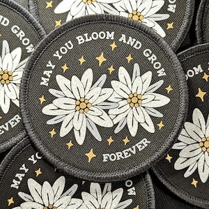 May include: Circular black patch featuring white daisy-like flowers and gold stars. The text "MAY YOU BLOOM AND GROW FOREVER" is printed around the edge. The patch has a stitched border.