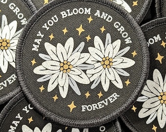Peut inclure: Écusson rond noir avec des fleurs blanches en forme de marguerite et des étoiles dorées. Le texte "MAY YOU BLOOM AND GROW FOREVER" est imprimé sur le bord. Bordure cousue.