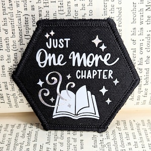 以下が含まれることがあります： 黒い六角形のワッペンで、白い文字で「JUST One more CHAPTER」と書いてあり、白い本のイラストが描かれていて、そこから魔法の渦巻きが出ています。