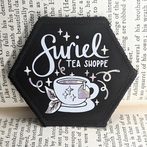 May include: A black hexagonal patch with white text that reads "Suriel Tea Shoppe". The patch features a white outline of a teacup with a tea bag and a leaf design. The teacup has a holographic finish.