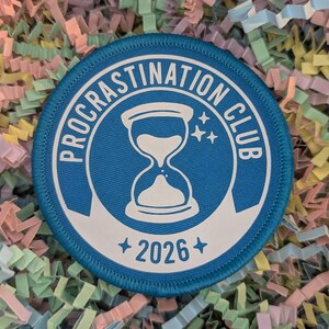 May include: A circular blue patch with white text, reading "PROCRATINATION CLUB" around the edge. An hourglass graphic and the text "2026" are in the centre. The patch is set against a background of colourful paper shreds.