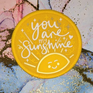 May include: A circular yellow patch with the text "you are my sunshine" in white script. The patch has a smiling sun graphic and star details. This patch is likely for clothing or accessories.