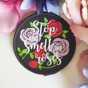 May include: Black round patch with white text that reads "Stop and smell the roses". The patch is decorated with pink, red, and green roses.