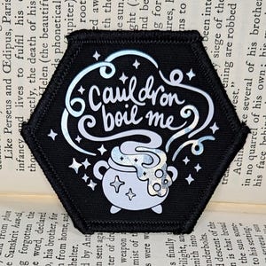 May include: A black hexagonal patch with a white cauldron and steam. The text "Cauldron boil me" is written in a white cursive font.