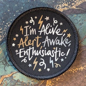 May include: A circular black patch with a stitched border featuring the white and gold text I'm Alive, Alert, Awake, Enthusiastic! The design is decorated with small stars, lightning bolts, and swirling line accents scattered across the surface.