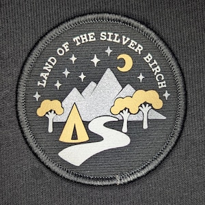 May include: A black and white embroidered patch with the text "Land of the Silver Birch" in a circle. The patch features a stylised image of a mountain range, a crescent moon, stars, a tent, and a winding path.