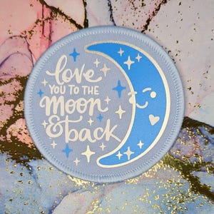 Może przedstawiać: Okrągła łatka w jasnoniebieskim kolorze z białym półksiężycem i gwiazdami. Tekst "love you to the moon & back" jest napisany białym, ozdobnym pismem.