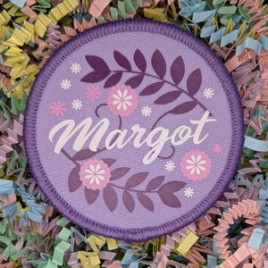 May include: Purple round fabric patch with a floral design and the name "Margot" in white lettering.
